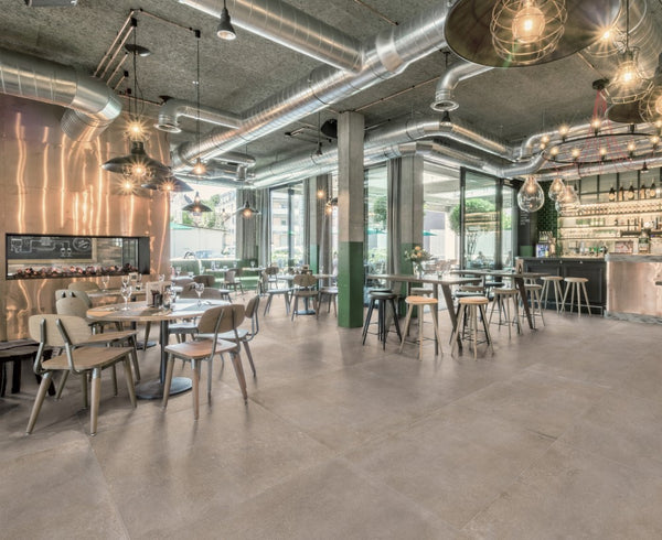 Slab tegels Durstone Taupe gelegd in een trendy restaurantinterieur met industriële accenten en natuurlijke verlichting door grote raampartijen.