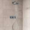 Moderne inbouw doucheset met ronde regendouche en handdouche in chroom, gemonteerd op een lichtgrijze tegelwand in een minimalistische badkamer, met stromend water zichtbaar.