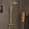 Thermostatische regendouche Brondby in geborsteld goud, gemonteerd in een moderne badkamer met donkergrijze wandtegels en bijpassende messing accessoires.