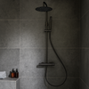 Thermostatische regendouche Brondby in geborsteld gun metal, geïnstalleerd in een moderne badkamer met donkere wandtegels en bijpassende accessoires.