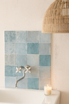 Wand met zellige tegels class zellige azul 10x10 cm achter wastafel met retro kraan, sfeervol verlicht door kaars en rotan lamp in natuurlijke badkamer.